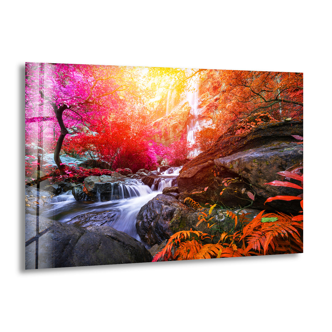 The "Waterfall in Forest View Tempered Glass Wall Art" showcases a vibrant fantasy landscape, featuring a cascading waterfall surrounded by colorful autumn foliage. The scene includes an array of red, orange, and pink trees with bright yellow sunlight filtering through the canopy, highlighting the flowing water and moss-covered rocks.