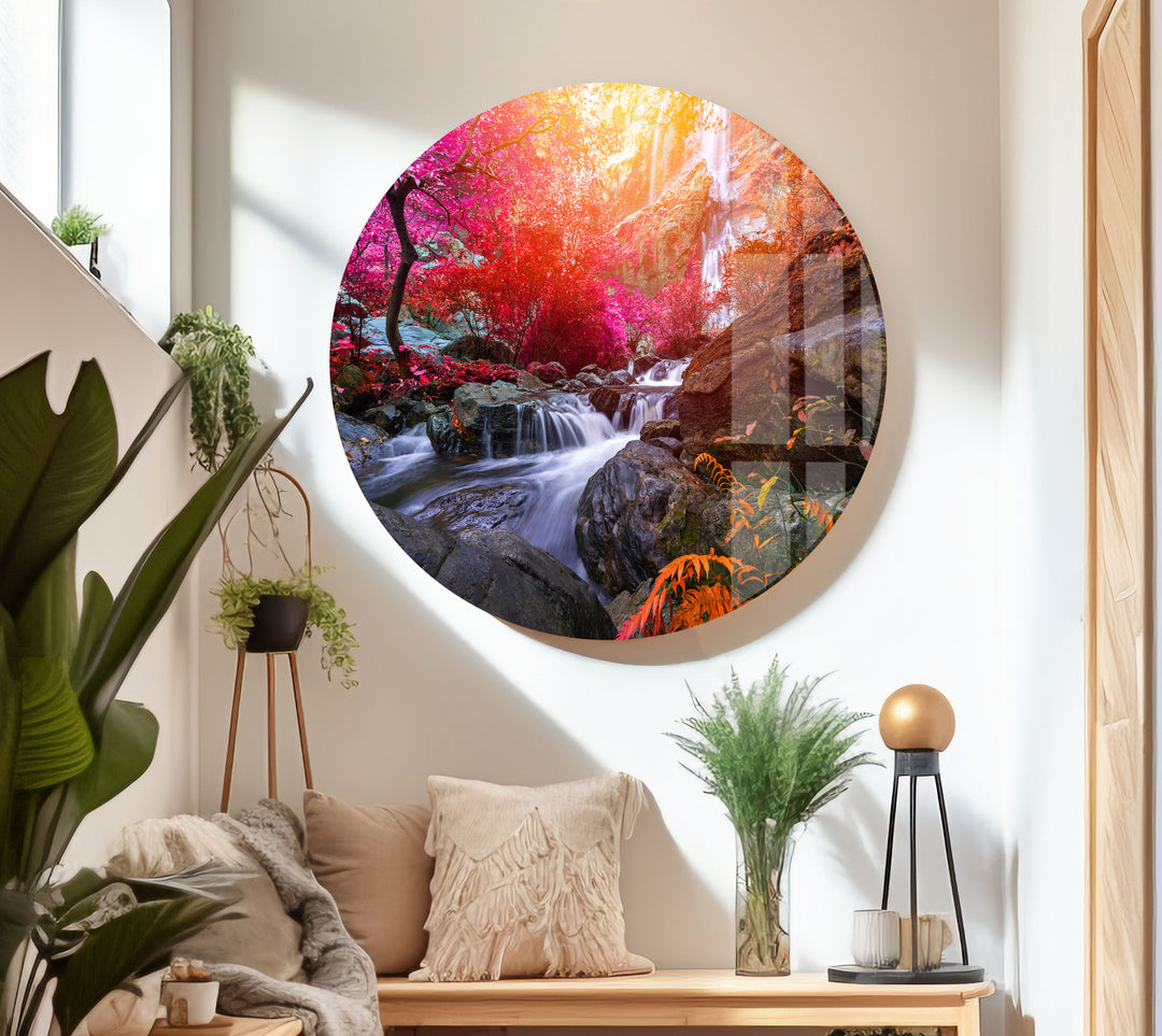 A cozy living room features the vibrant Waterfall in Forest View Tempered Glass Wall Art, which beautifully depicts a colorful forest with a waterfall. Surrounded by plants, the space includes a beige couch adorned with cushions, a small plant stand, and a modern lamp on a white table. Natural light floods the room, enhancing its interior decoration.