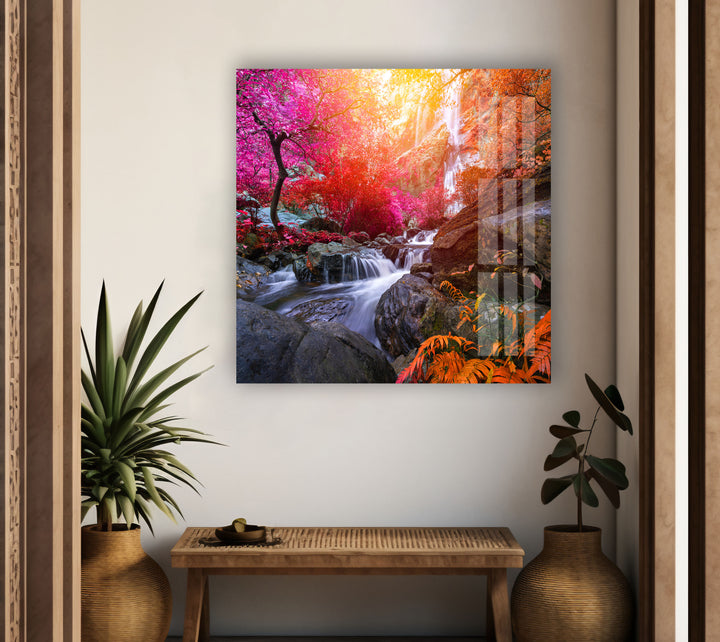 A brightly lit room with a wooden bench and two plants on either side. A large, colorful piece of art titled "Waterfall in Forest View Tempered Glass Wall Art," featuring a vibrant autumn forest with a waterfall and rocky stream created using UV printing technology, is hung on the white wall, adding a splash of color to the serene and minimalist interior.