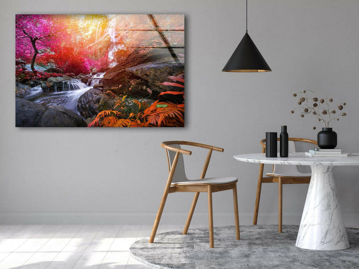 A minimalist dining area features a round marble table with a white top, surrounded by two wooden chairs. A black pendant lamp hangs above. The wall showcases the Waterfall in Forest View Tempered Glass Wall Art, depicting a waterfall in a colorful forest using UV printing technology. Dried flowers and black vases decorate the table, adding charm to the interior decoration.