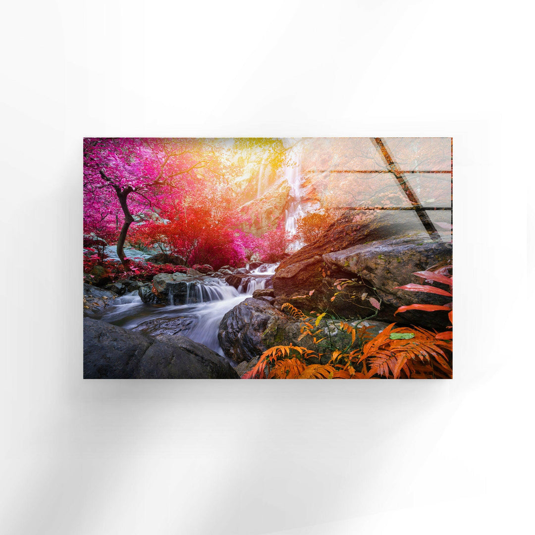 An open book featuring a vibrant, colorful image of a serene waterfall cascading over rocks, surrounded by lush, multi-colored foliage—including pink and red trees and orange ferns—appears to float against a simple white background. This captivating scene could be the perfect inspiration for your next piece of Waterfall in Forest View Tempered Glass Wall Art using UV printing technology, ideal for interior decoration.