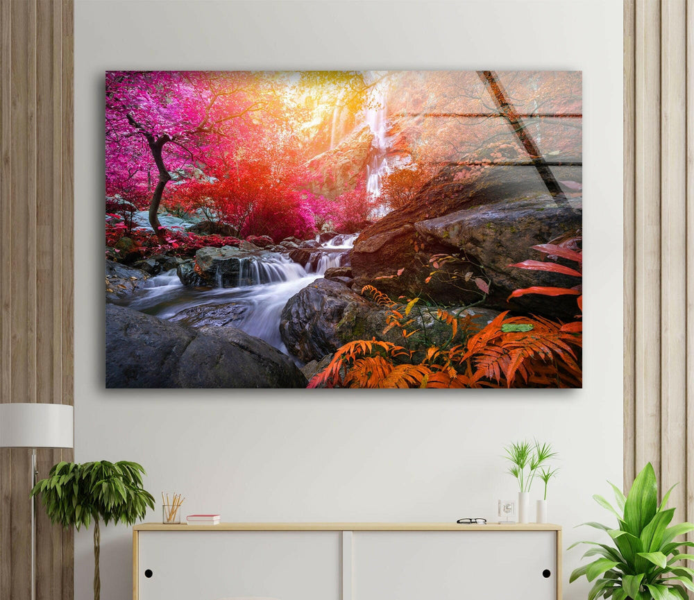 A large, vibrant photograph named "Waterfall in Forest View Tempered Glass Wall Art" is mounted above a minimalist desk. The wall, flanked by wooden panels, enhances interior decoration with small plants, a lamp, books, and a cup adorning the desk.