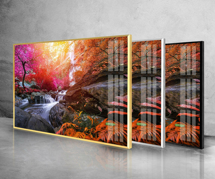 Three framed images are displayed in a staggered arrangement. The primary piece, the "Waterfall in Forest View Tempered Glass Wall Art," features a vibrant autumn scene captured with UV printing technology, showcasing a flowing waterfall amid colorful foliage and rocks. Perfect for interior decoration, the gold, white, and black frames cast reflections on the shiny surface below.