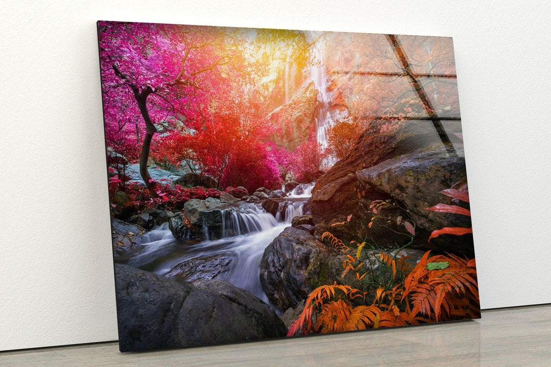 Introducing the Waterfall in Forest View Tempered Glass Wall Art. This captivating piece features a vibrant, colorful forest scene where a waterfall cascades gracefully over rocks. The trees, dressed in bright pink, red, and orange foliage, create an ethereal atmosphere. Utilizing UV printing technology for vivid colors, this artwork appears stunning against a white wall on a wooden floor.